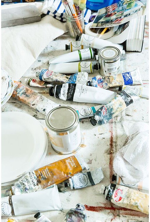 Tubes of paint and glass jars filled with brushes and tools cover a table in Mary’s light-filled studio.
