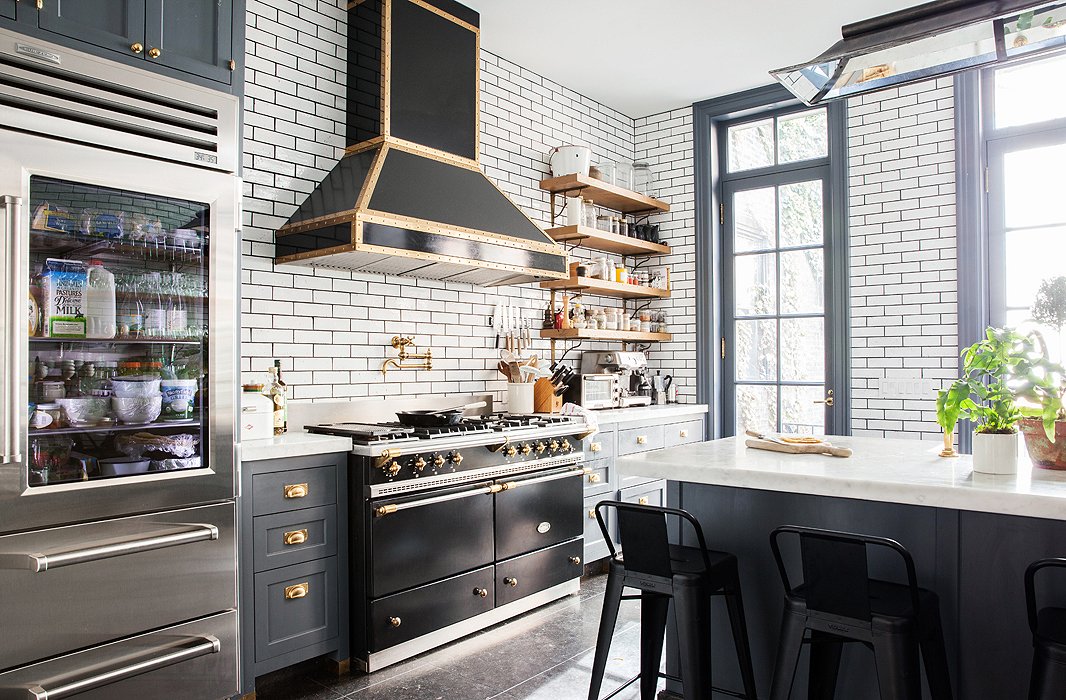 A black Lacanche stove is a clear centerpiece of the kitchen. The walls are covered in a bricklike subway tile, framed by dark grout.
