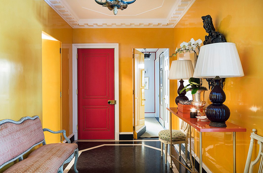 Kate wanted the windowless foyer to be both warm and inviting. The solution? “Guts and drama” by way of lacquered walls (painted in Farrow & Ball’s Orangery), stone floors, and an Eve Kaplan mirror. The pièce de résistance is a door upholstered in red Dualoy leather embellished with brass nail heads. 
