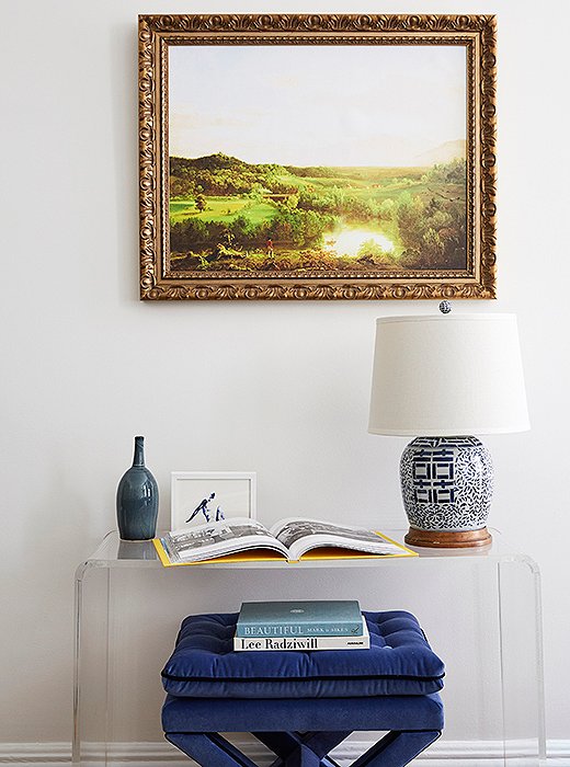 Opposite the fireplace, an acrylic console (one of the few pieces brought from Amy’s previous apartment) offers a spot to display favorite photos, books, and objets. A tufted ottoman tucked underneath adds a layered look—and handy seating.
