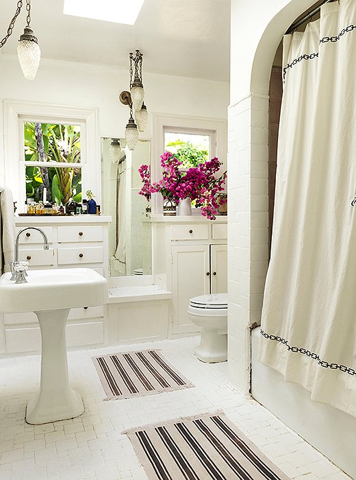 The bath has original built-ins and Batchelder tile. “I painted it all white,” says Heidi. “People gasp, ‘You painted it? That tile is iconic!’ But it was brown, and I didn’t like it. It is still there.” Heidi found Moroccan-style pendants to go with the mats and the embroidered shower curtain. Bougainvilleas from the garden fill a rustic pitcher.
