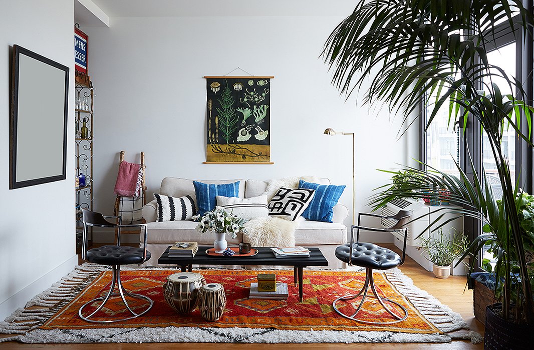 The apartment’s living room illustrates the home’s colorful, collected aesthetic, with a German teaching scroll, lush layered rugs, mud-cloth pillows, and a pair of midcentury swivel chairs.
