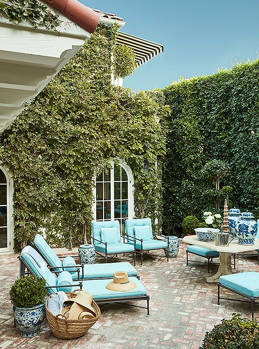 A courtyard, framed by ficus hedges, makes a lush spot for relaxing or dining year-round. “I love this antique stone table, even though it’s crumbling, and designed the entire space around it,” Mark says. “We use it for dining, as a bar, or just as a focal point.” A straw tote is filled with sun hats; garden stools are used as both side tables and seating.

