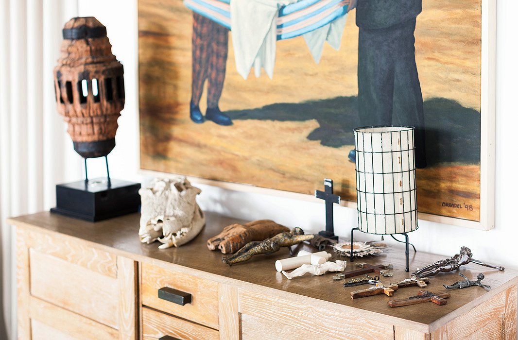 A collection of religious crosses is displayed alongside a sculptural lamp atop a lightly weathered 1940s French wood cabinet in the bedroom.
