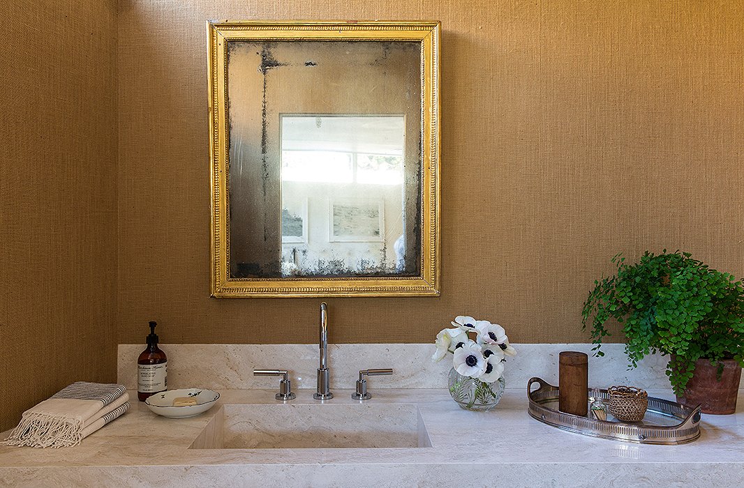 A cool travertine vanity is a perfect foil to the 19th-century mirror. The color throughout the home is Benjamin Moore’s White Dove, but for this space, Jessica chose burlap wallpaper. Anemones and maidenhair ferns are two of the couple’s favorite plants.

