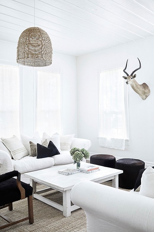 A taxidermy bust stands watch over the Holly House living room, which features crisp white linen upholstery, dark hair-on-hide ottomans, and a mix of striped and Anatolian kilim pillows. 
