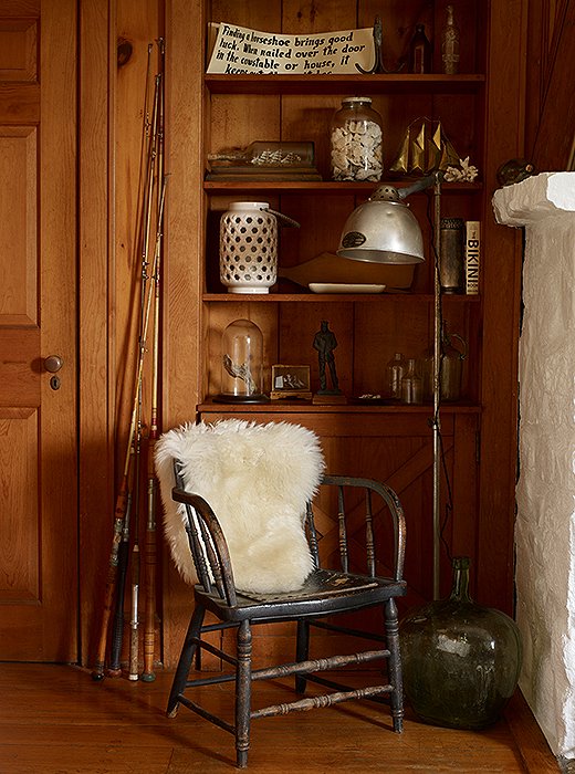 A captain’s chair draped with a sheepskin creates a cozy corner next to the living room’s fireplace. Behind, a built-in bookshelf lined with thrifted treasures reads as an open-air cabinet of curiosities. 
