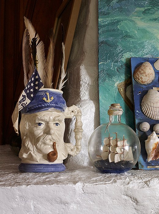 A stone mantel painted white hosts a ship in a bottle, a novelty mug, and artwork whose watery colors mirror the surface of the sea. 
