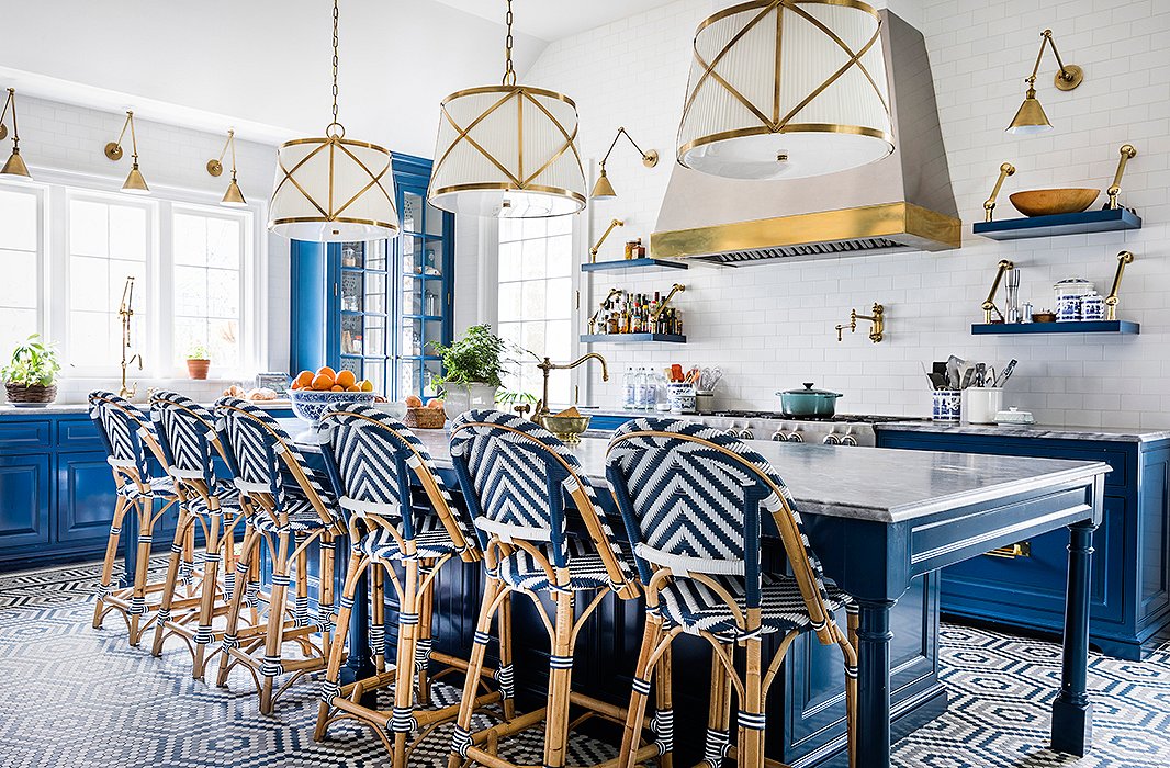 What was once a garage is now the kitchen. Hexagonal encaustic-tile floors draw from the hues of the cabinetry and the Gris de Savoie marble countertops. Brass pendants, sconces, and an exposed-steel range hood add reflective flair, while blue-and-white bistro stools encourage guests to pour a cup and stay awhile. Find similar sconces here.
