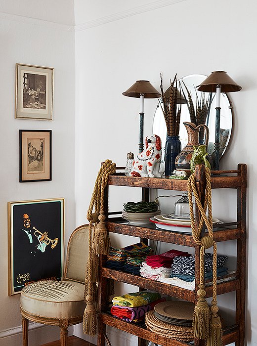 A cart draped with tassels hosts napkins, not liquor. On top, a Staffordshire dog holds court between two buffet lamps in front of pheasant plumes and a vintage mirror.
