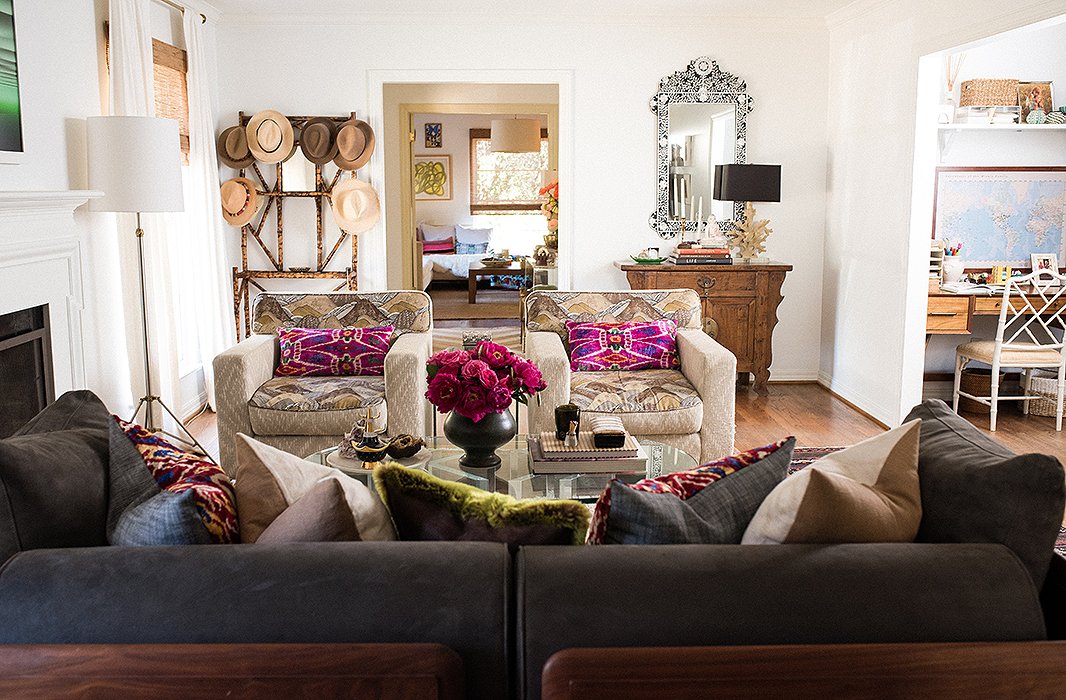 Vibrant Gaia pillows accent a pair of neutral club chairs in the living room. A bamboo hall tree and a bone-inlay mirror frame the doorway behind.

