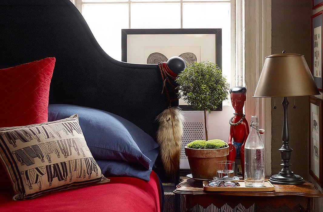 In the bedroom, an iron bed from Ralph Lauren Home is made with sheets that match the hue of the kitchen shelves. At bedside, an African statue stands guard just behind a cork tray holding spectacles, a glass (half full), and a small topiary. 
