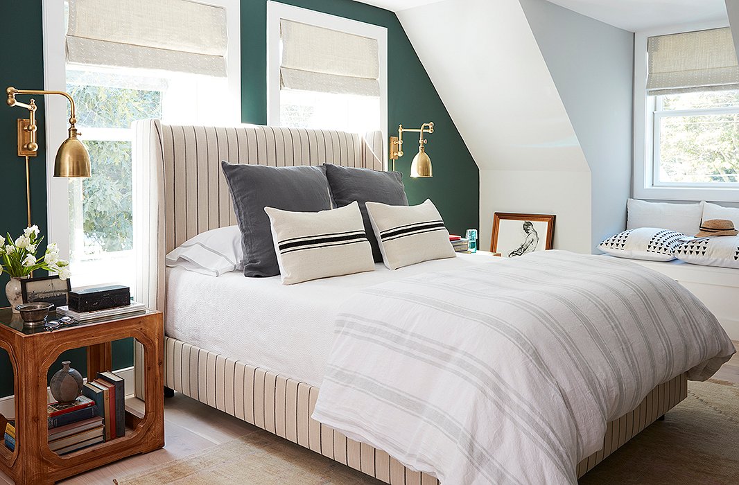 In the master, an upholstered bed was placed in front of double-hung windows to maximize the room’s layout. Brass swing-arm sconces lend the arrangement a sense of place and cast an unobtrusive downward glow when on. 
