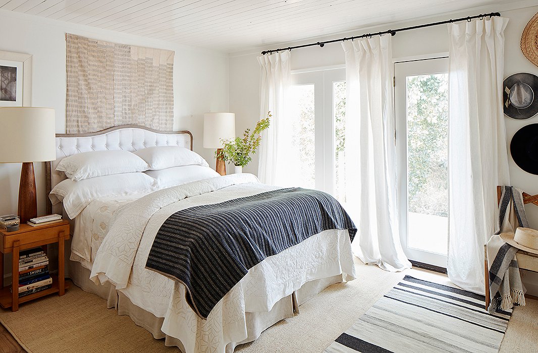 In the master bedroom, an upholstered headboard by Blink is flanked by vintage Scandinavian lamps and Saarinen side tables. Behind, an African textile adds a textural layer of interest and reflects the tones of the room’s natural-fiber rug and striped runner. 
