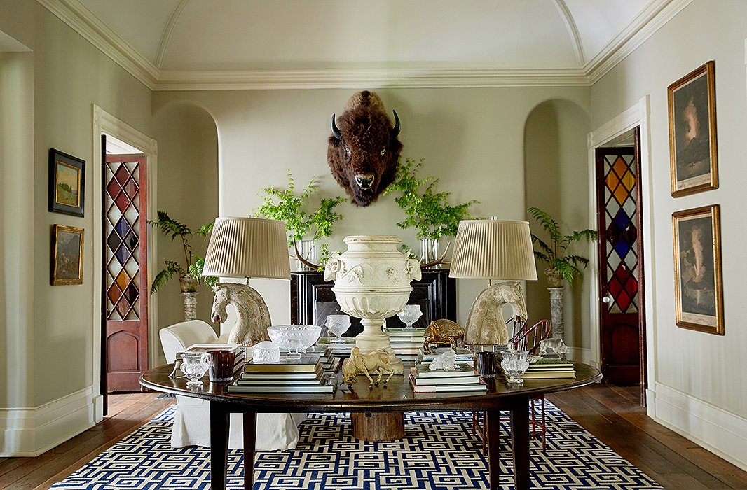 In the foyer, most all is vintage save the blue-and-white rug emblazoned with Greek keys. From between branches (cut from the yard) hangs Otis the bison, watching over a central table stacked with coffee table books and Dale’s collection of Lalique glassware. Sculpted stallions crowned by pleated shades flank a marble urn, emphasizing the home’s ties to horse country. 
