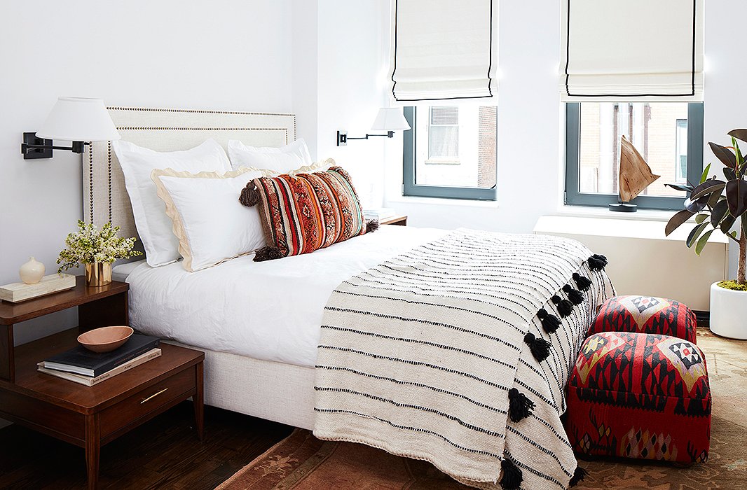 Textiles carry the eye in the white-walled master bedroom. “I really learned from my past decorating mistakes,” says Mindy. “I would fall in love with a really bright specific color, end up painting the whole room that color, and get really sick of it.” Catastrophe, avoided. On the windows, Roman shades from The Shade Store continue a theme of black details with stitched borders, tying into the tasseled quilt and bronze accents. 
