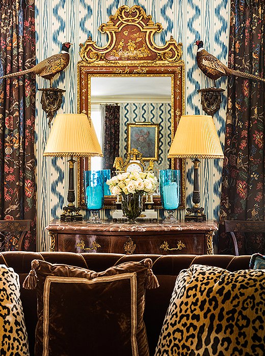 An English chinoiserie mirror was hung above a Louis XV-style commode and flanked with a pair of pheasants. Touches of gold echo the glow of the sofa’s brown velvet as it catches the sun. 
