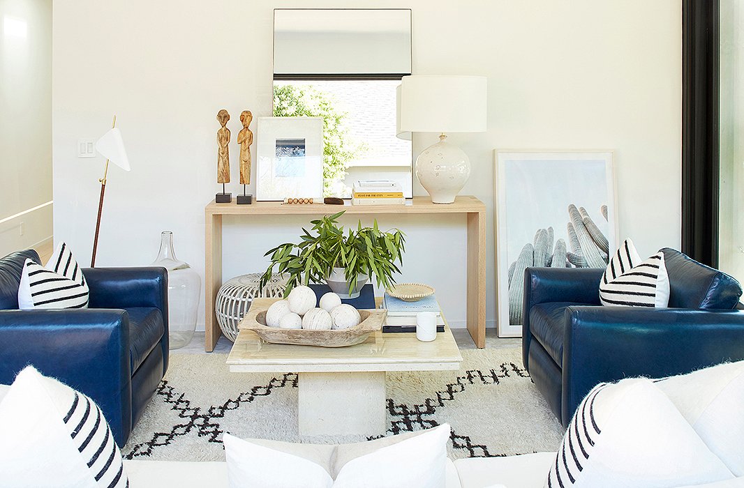 Between a midcentury-style floor lamp by AERIN and a cacti print casually leaned against the wall sits a console arranged with a ceramic lamp, an oversize mirror, and a white rattan Albini ottoman. 
