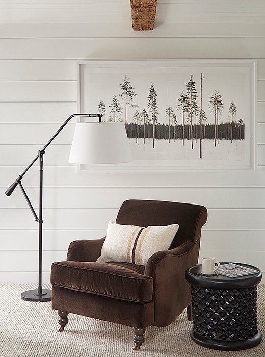 A chair upholstered in rich brown velvet sits next to a floor lamp by Ralph Lauren Home and a carved-wood stool turned side table.
