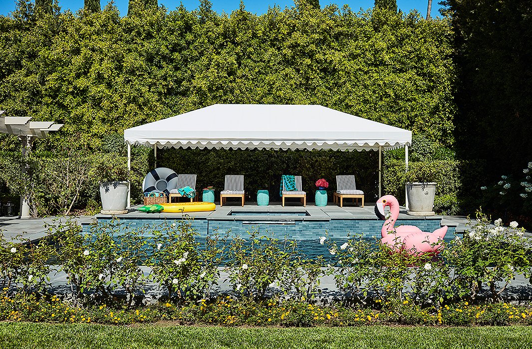 Poolside, a striped and scalloped cabana provides shade for a row of loungers. A flamingo by SunnyLife drifts close by. 
