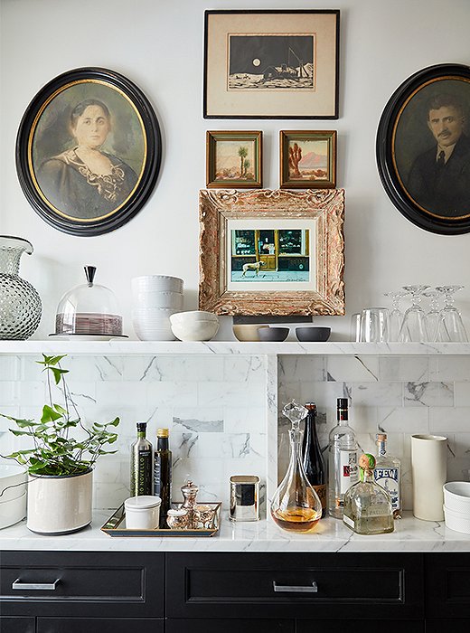 A medley of vintage artworks lends spirit to the black-and-white kitchen. The slim marble countertop and shelf house a tightly curated collection of barware, kitchen accessories, and serving pieces.

