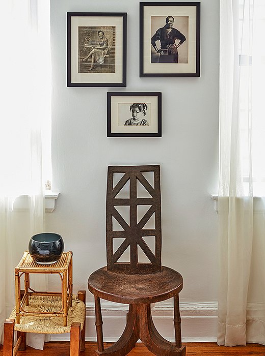 A trio of black-and-white photographs hang above a carved wooden chair, next to which stand two stools, artfully stacked to serve as a side table. 
 
