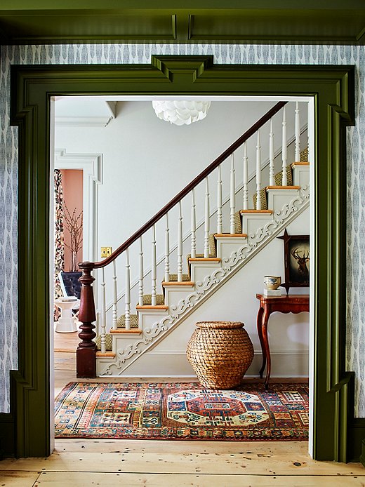 Farrow & Ball‘s Bancha paint color on the molding creates a striking green contrast to the rest of the living room. The entryway features delicate woodwork along the staircase. The vintage runner is part of Hendrick Churchill’s collection of heirloom rugs. 
