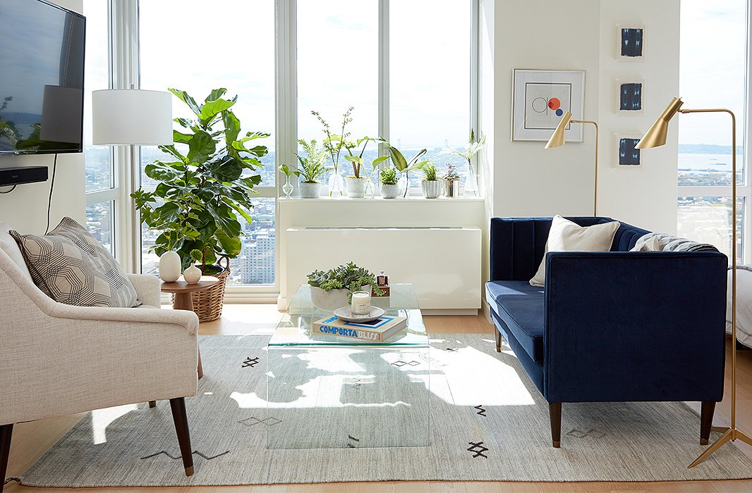 A glass coffee table offers a useful surface without hindering light or a sense of space. The velvet settee and the brass lamps visually separate the seating area from the sleeping alcove, while the rug speaks to the neutral hues found throughout the apartment. 
