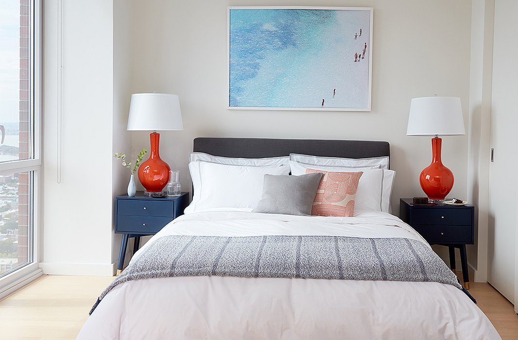 Blue nightstands were selected in the interest of color and storage, pairing perfectly with orange lamps and the oversize aerial photograph hanging above the bed upholstered in charcoal linen. 
