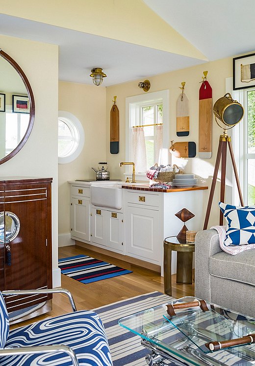 The guesthouse’s petite kitchen is home to an apron-front sink, cutting boards painted in a nautical palette hung on the wall, a gauzy window covering from Schumacher, and brass fixtures. 
