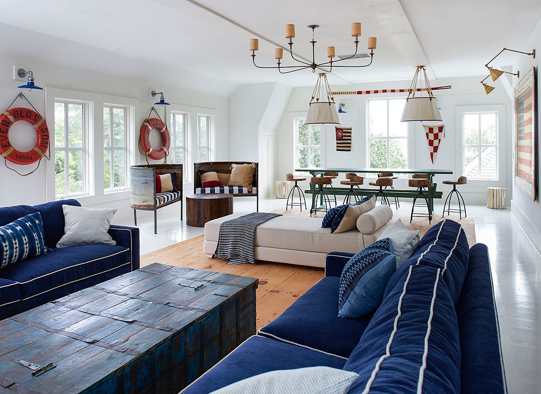 The party barn, which sits above the property’s garage, is the perfect place for the clients’ kids and friends to hang out and relax. Ralph Lauren Home’s Miramar Hanging Shades sit above the vintage dining table, which is surrounded by a set of swivel chairs by Arteriors. The vintage life rings and mud-cloth textiles are from the Brimfield Antiques Show, and the pair of chairs made from a reclaimed trough are from the Round Top Antiques Show. 
