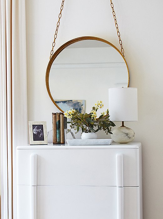 “I always adore a hanging mirror,” Caitlin says, and the brass cord of this one plays off the brass of the bedside mirrors. The dresser, a midcentury piece coated in white lacquer, perfectly complements the rest of the room—and gives her husband enough space for his clothes. 
