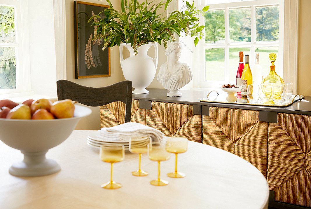 Sea-grass doors in a classic rush weave bring organic allure to an otherwise simple mahogany sideboard. Elements such as the amphora-inspired vase and the classical bust atop the sideboard, the pedestal bowl on the table, and the botanical art on the wall show how beautifully Old World elegance complements earthy textures.
