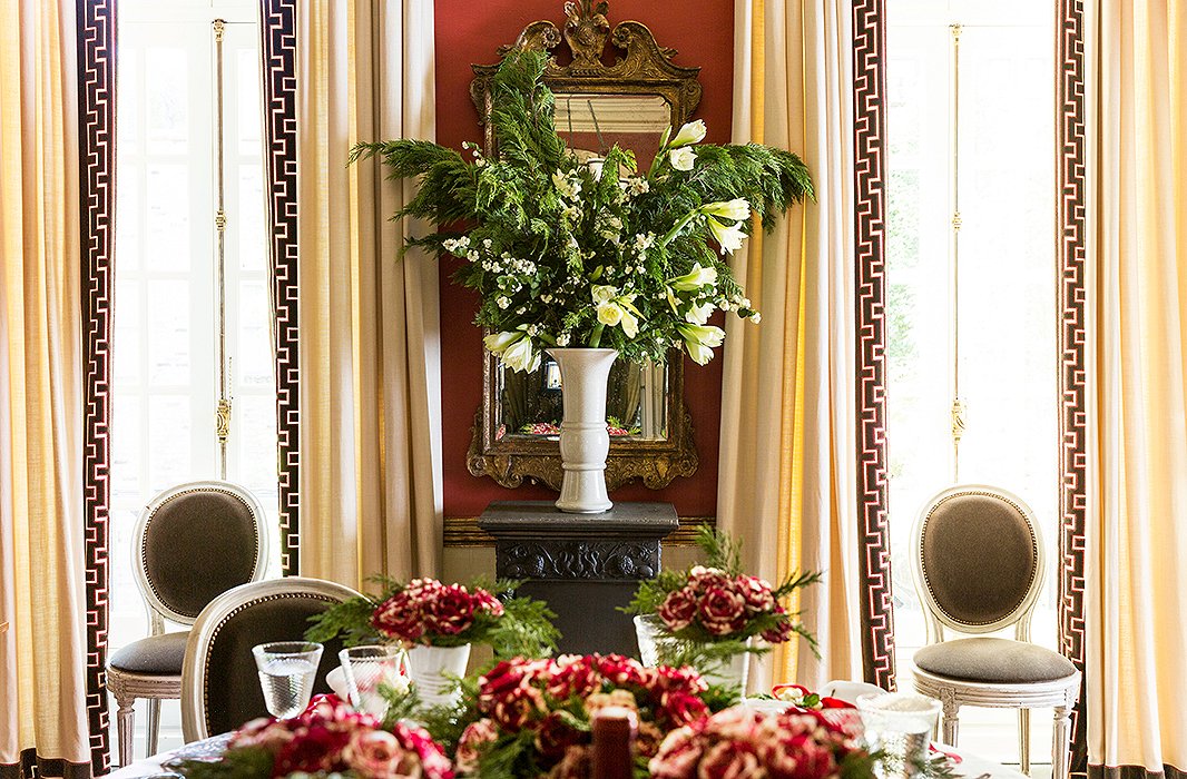 Leafy greenery overflow on the table, while a blooming vase of pine branches, winter greens, amaryllises, and white berries stands in for a tree and steals the spotlight as a floral centerpiece.
