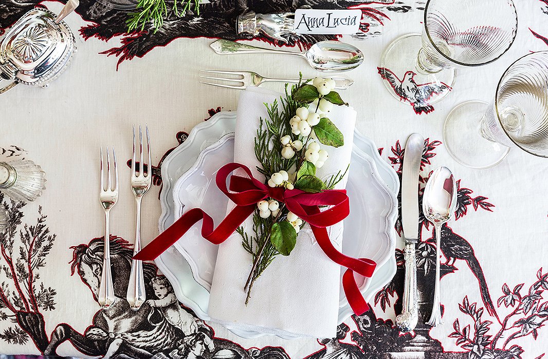 The place settings feature dishes by Astier de Villatte, embroidered napkins, and 18th- and 19th-century silver; a velvet bow and a sprig of pine and white berries is in keeping with Alessandra’s festive palette.
