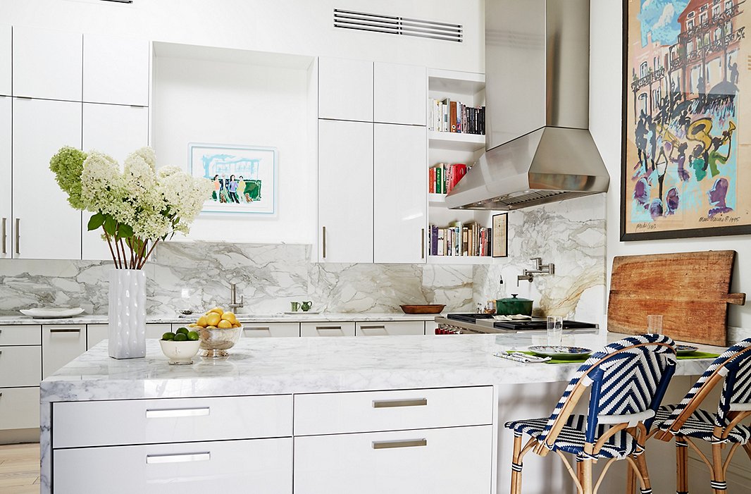 Framed artwork, striped bistro stools, and colorful cookbooks on open shelves add dimension to the all-white kitchen.
