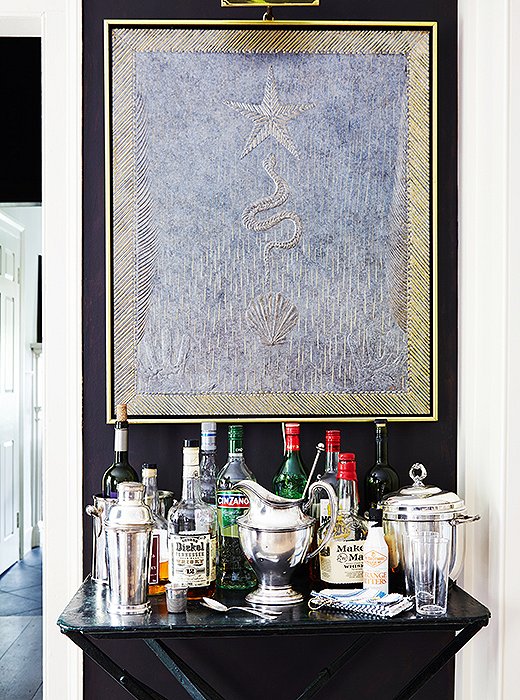 Dark walls and a dark marble-top table make a dramatic contrast to glittering barware and one of Frank’s luminous gilt-edged paintings.
