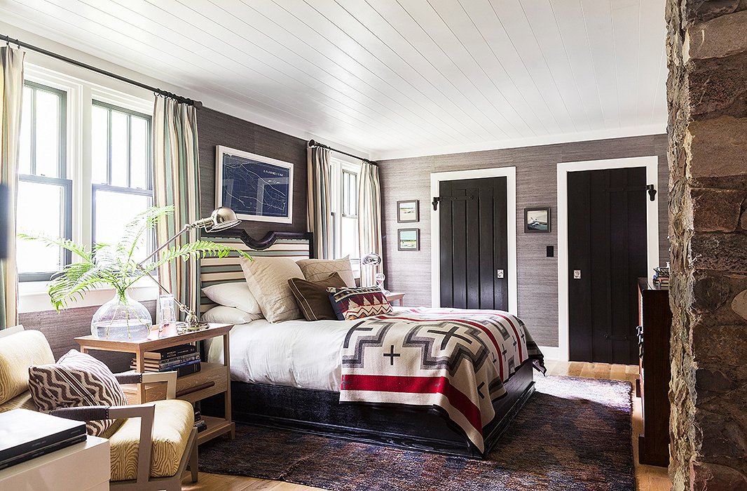 Fabric-covered walls, an upholstered headboard, and matching drapes make a sophisticated contrast to outdoorsy touches including a Pendleton blanket, a framed map, and dreamy seascapes.
